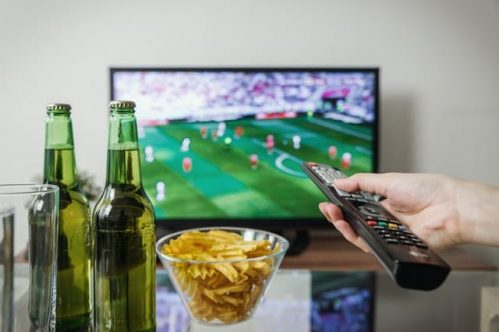 hand with remote pointing at television showing sports while there are snacks and drinks beside